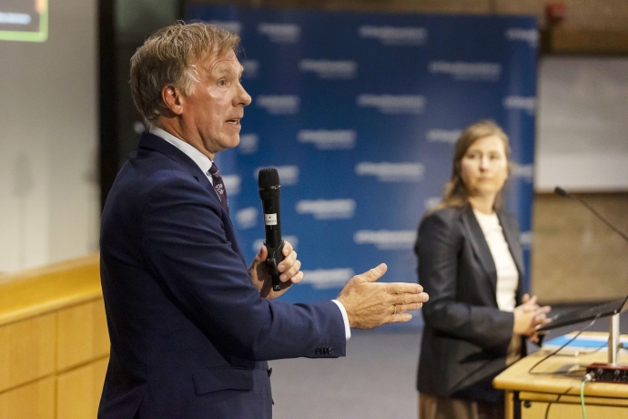 man Dr. Hommes in blue suit speaks holding microphone on stage as woman speaker with long dark hair looks on in background