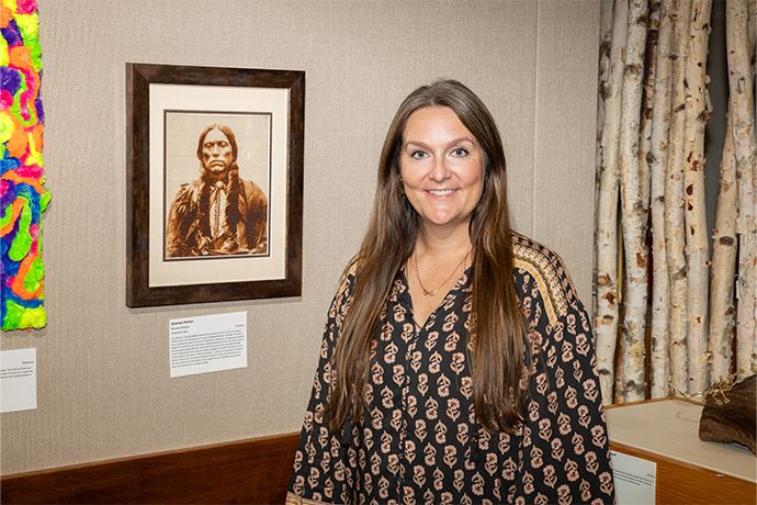 woman with long brown hair and patterned shirt stands in front of her artwork of Native American chief displayed on the wall behind her