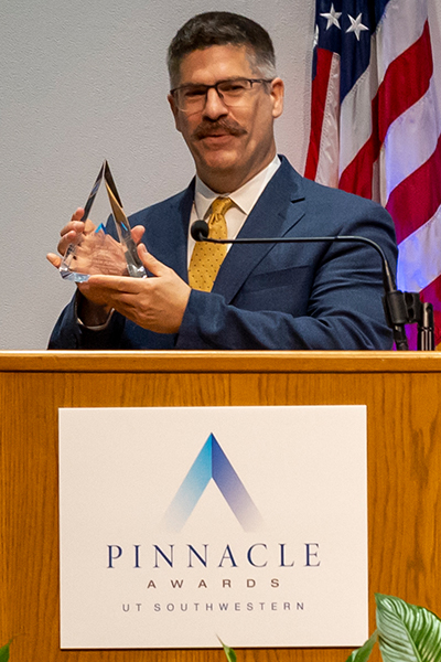 man with short dark hair and mustache holds glass trophy as he stands behind Pinnacle Awards podium, US flag in background