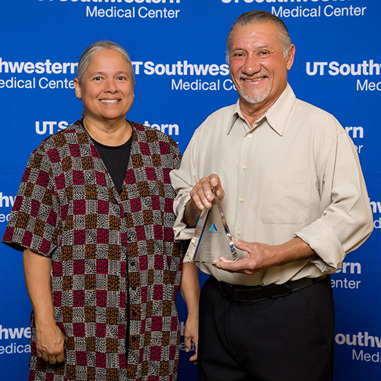 middle-aged woman and man in tan dress shirt holding glass trophy stand in front of UTSW backdrop