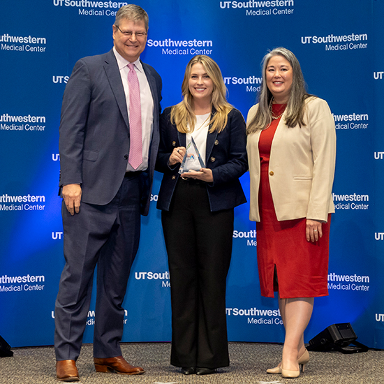 tall man in suit stands with 2 women, center woman holds glass trophy, in front of UTSW backdrop