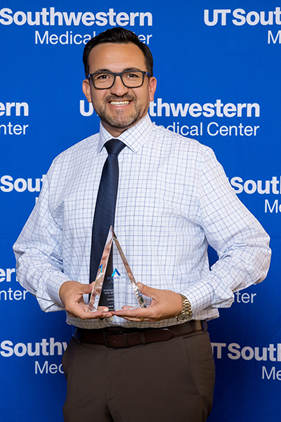 smiling man in white patterned dress shirt and dark tie holds glass trophy in front of UTSW backdrop