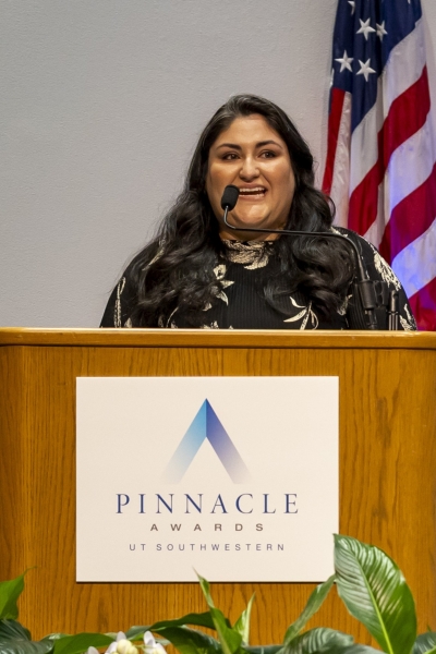 woman with long dark hair speaks at Pinnacle Awards podium, US flag in background