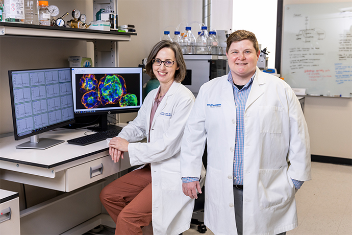 2 researchers in white lab coats stand in front of computer monitor in lab