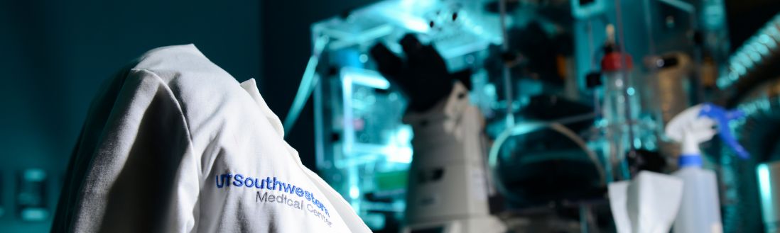 White lab coat with UT Southwestern medical school logo on chair, microscope on lab table in background.