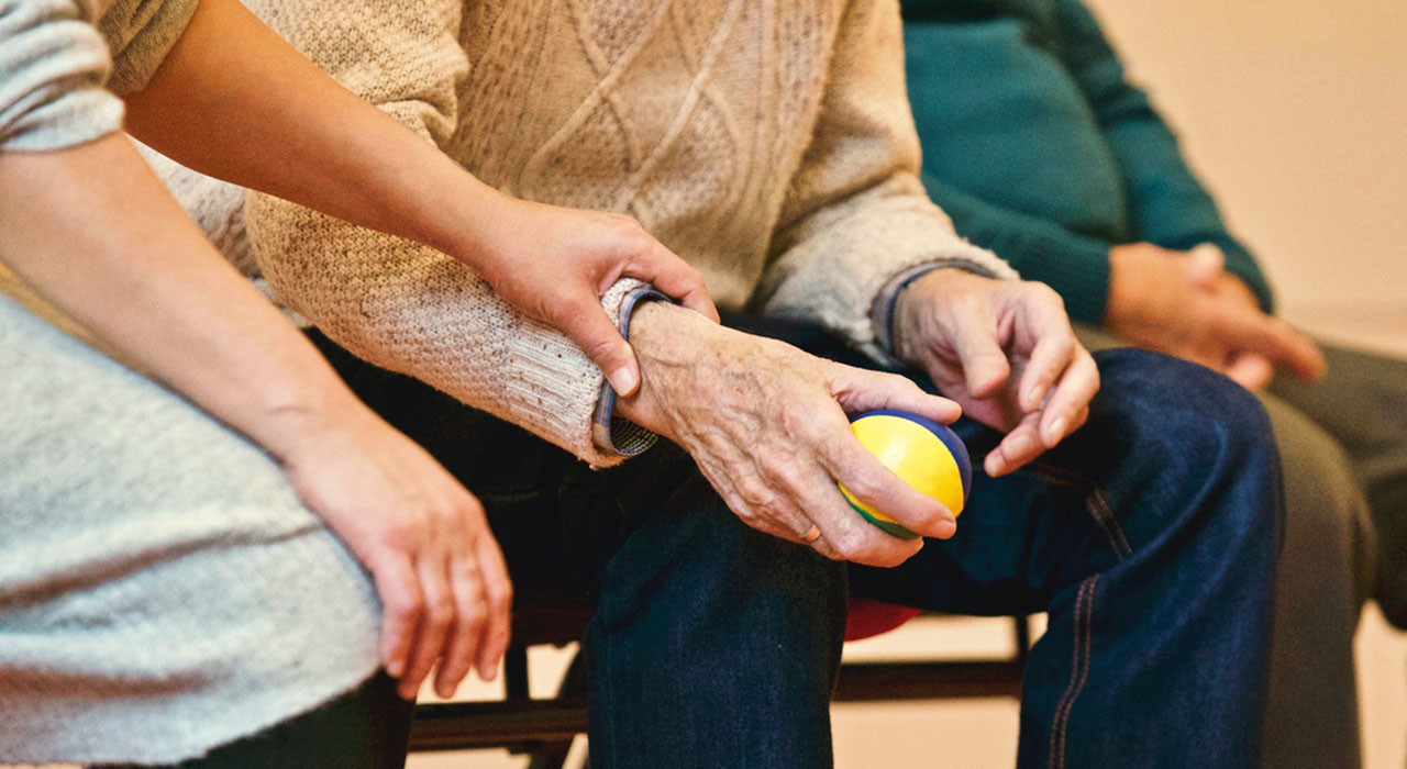 A close-up photograph of the hands of an elderly person holding a blue and yellow ball, as the hands of a physical therapist guides them through exercises