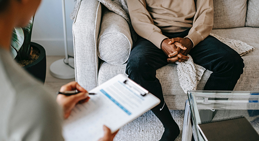 A closeup of a patient sitting with their hands clasped as a doctor writes on a clipboard