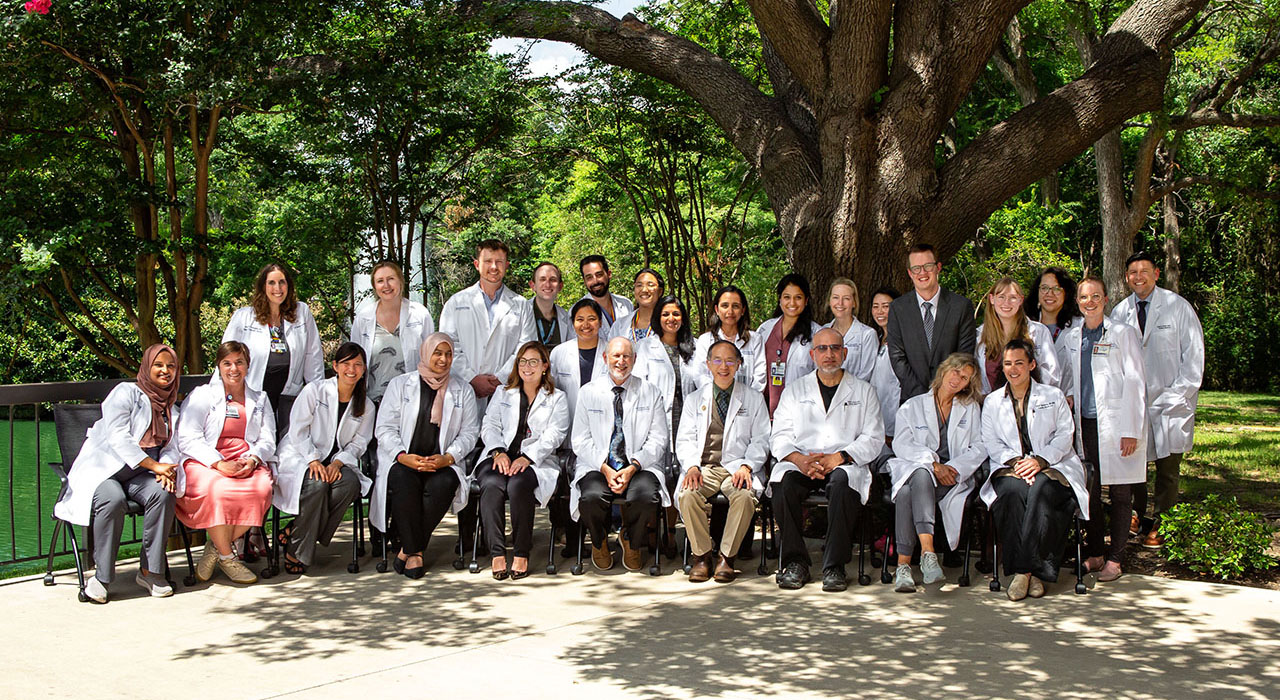 A group of smiling men and women wearing white lab coats, posed in front of a large tree.