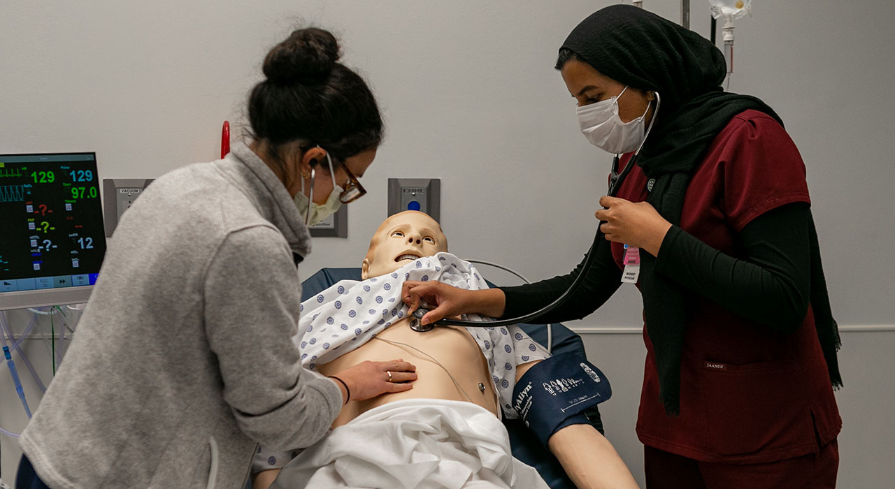 Two women wearing surgical masks perform a medical examinaiton on a training dummy