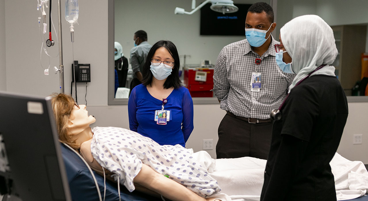 Three people wearing surgical masks look at a medical training dummy, which is looking at the ceiling