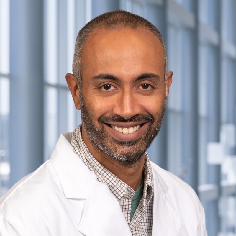 Dr. Kartik Rajagopaan wearing a checkered shirt and a white lab coat standing in the Clements University Hospital lobby