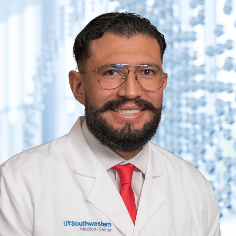 Dr. Lisandro Maya Ramos wearing a white shirt with a red necktie and a white lab coat standing in the Clements University Hospital lobby