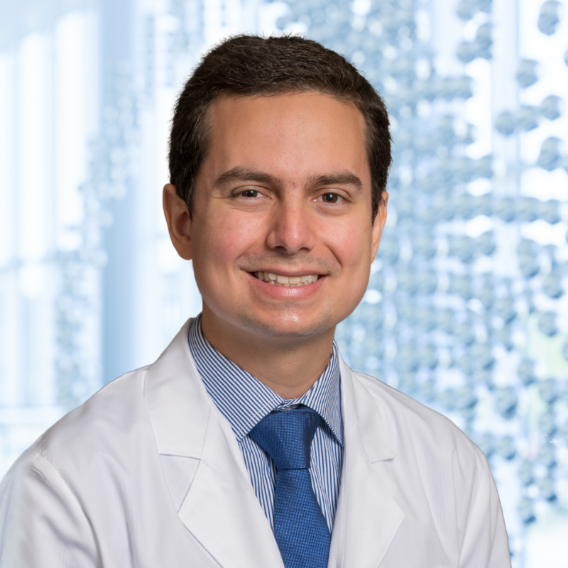 Dr. Juan Salcedo Betancourt wearing a blue stripped shirt with a blue patterned necktie, a white lab coat, standing in the Clements University Hospital lobby