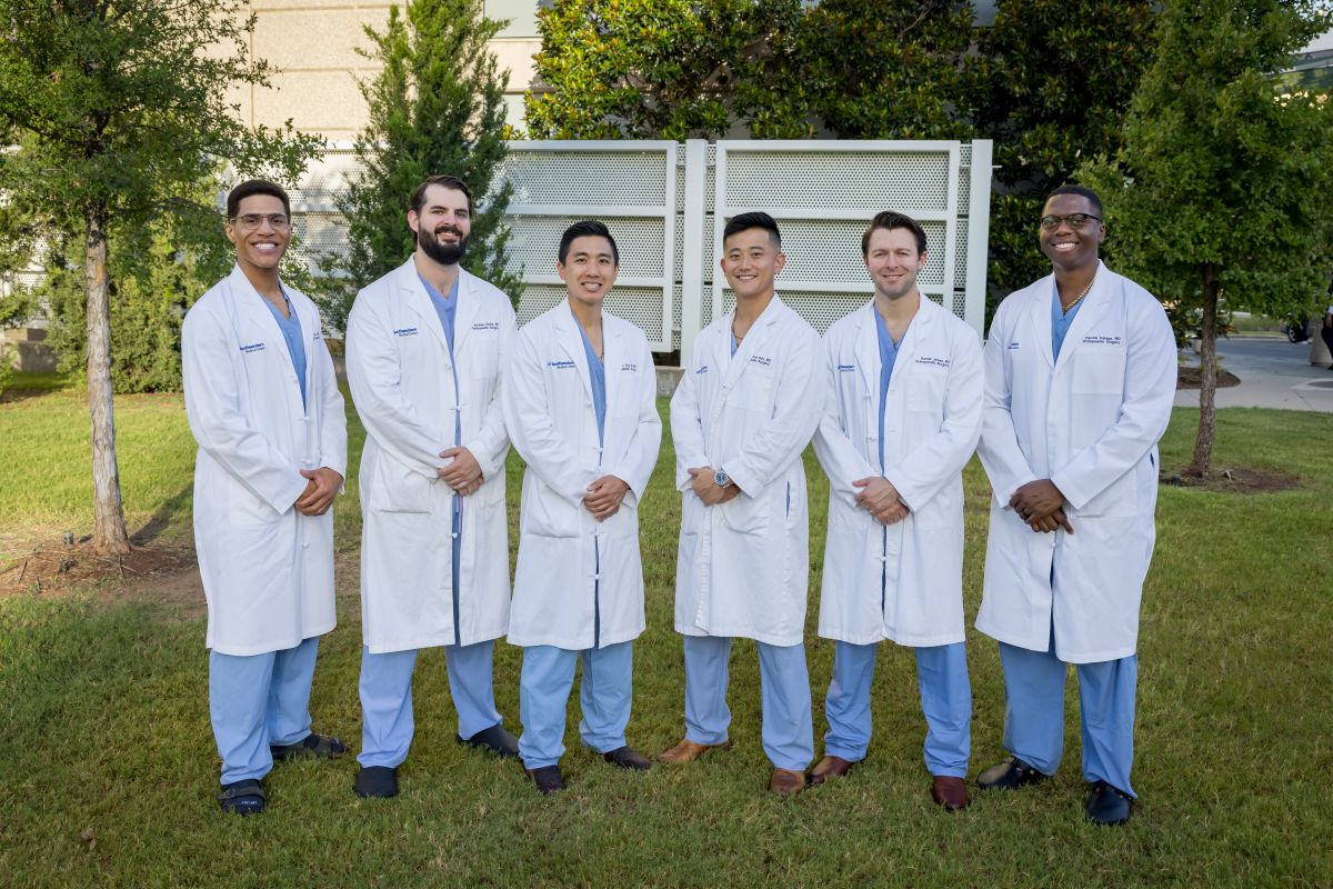 group of 4 men and 2 women Orthopaedic Surgery Residents wearing white coats and blue scrubs standing outside hospital