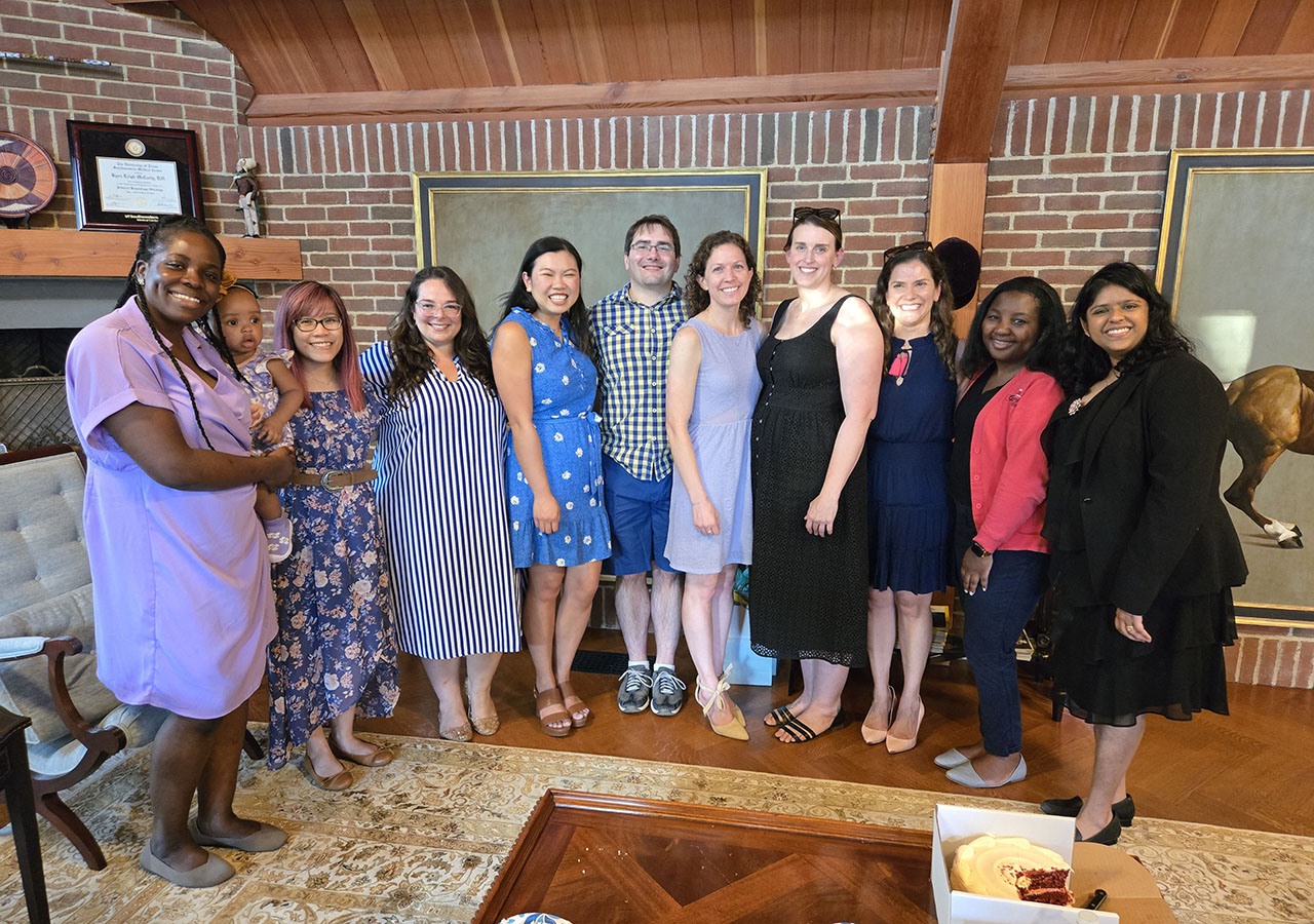 A group shot of the Fellows at a party with a cake in the foreground. The woman at left is holding a baby.
