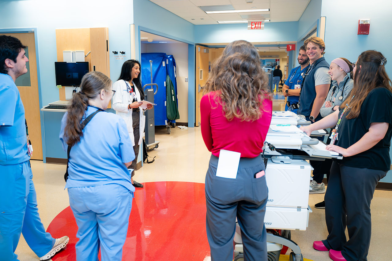 A woman in a white coat speaks to a group of students in a hospital hallway