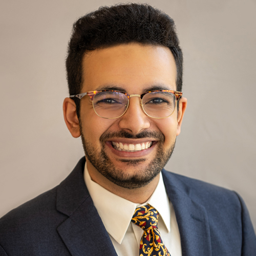 Dr. Harirah headshot, smiling man with curly dark hair, beard and glasses in suit and patterned tie