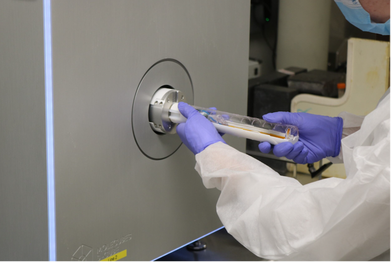 An individual in protective clothing, including gloves and a mask, handles a cylindrical object near a circular opening of a large metallic Molecubes imaging machine.