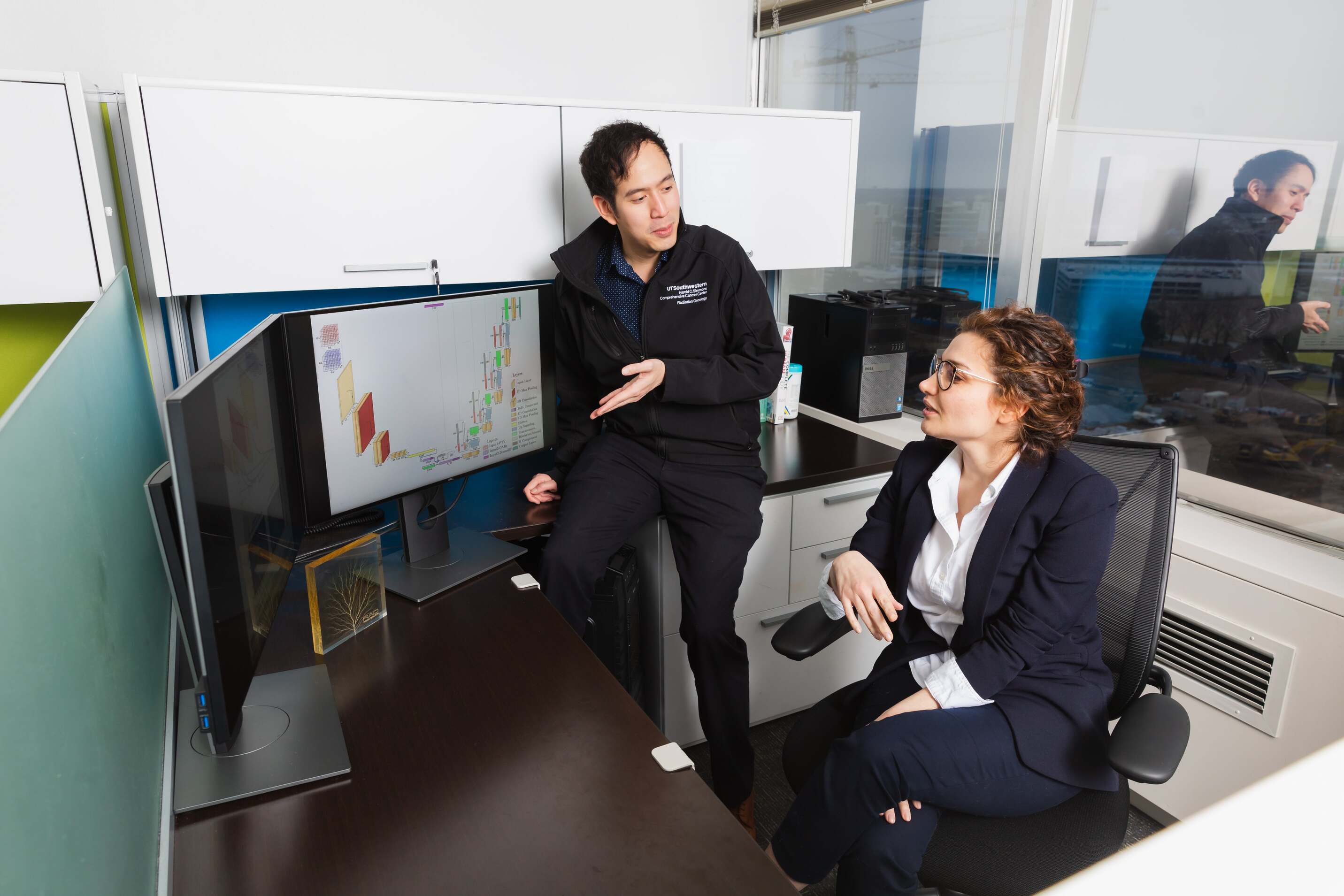 Two individuals in an office setting are reviewing a diagram displayed on a computer monitor, with one pointing at the screen and the other observing attentively.