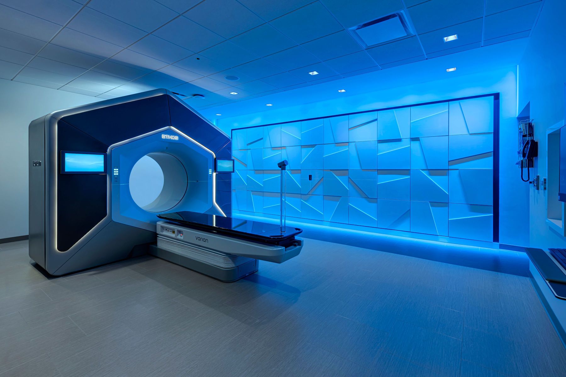 Modern radiation therapy room with blue ambient lighting, featuring a large ETHOS machine with a circular opening and a patient table, surrounded by control panels and geometric wall designs.