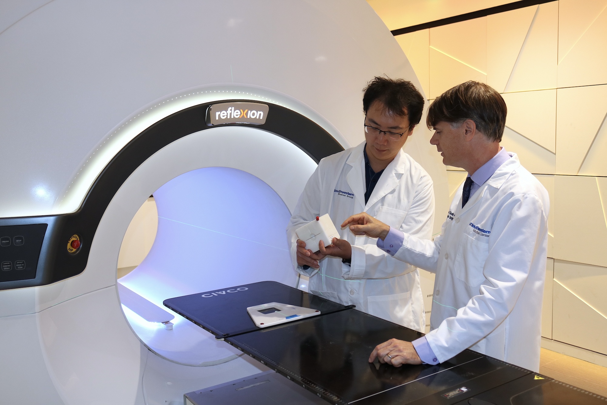 Two medical professionals in lab coats stand beside a RefleXion imaging machine.