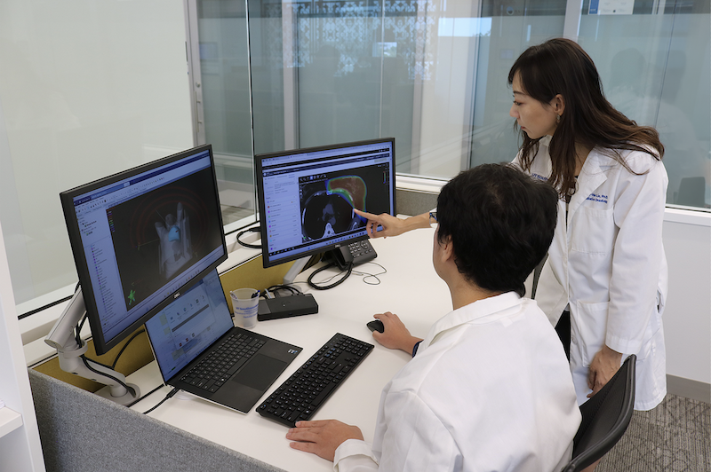 Two individuals in lab coats review cross-sectional medical scans on dual monitors; one is seated using a mouse while the other stands and points at the screen.