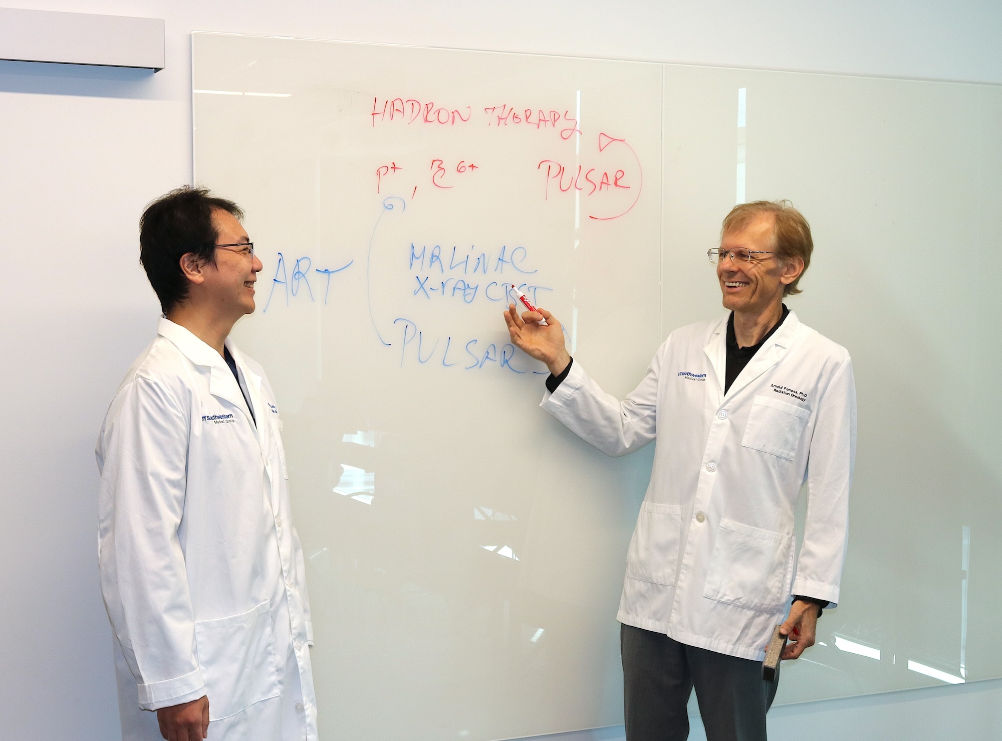 Two doctors in lab coats stand in front of a whiteboard; one points at the board, which displays terms related to radiation therapy.
