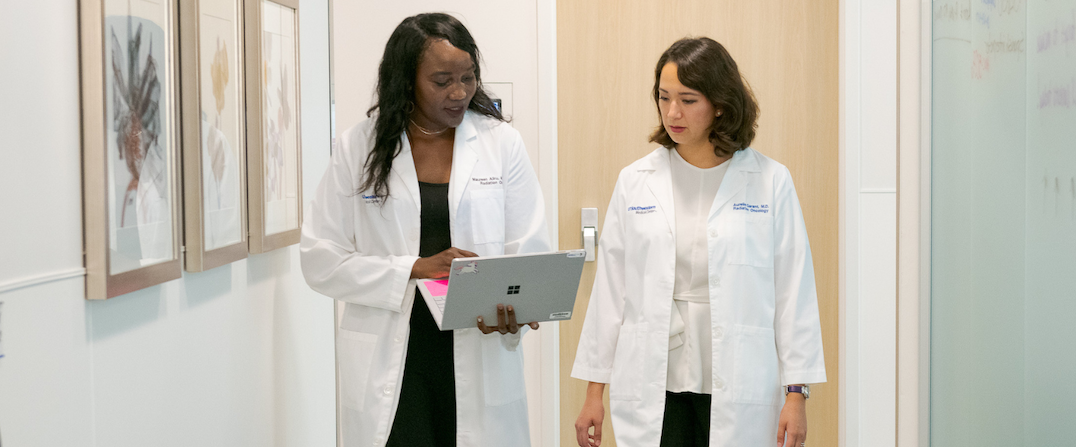 Two medical professionals in white lab coats walk down a hallway lined with framed pictures; one holds a tablet while conversing with the other.