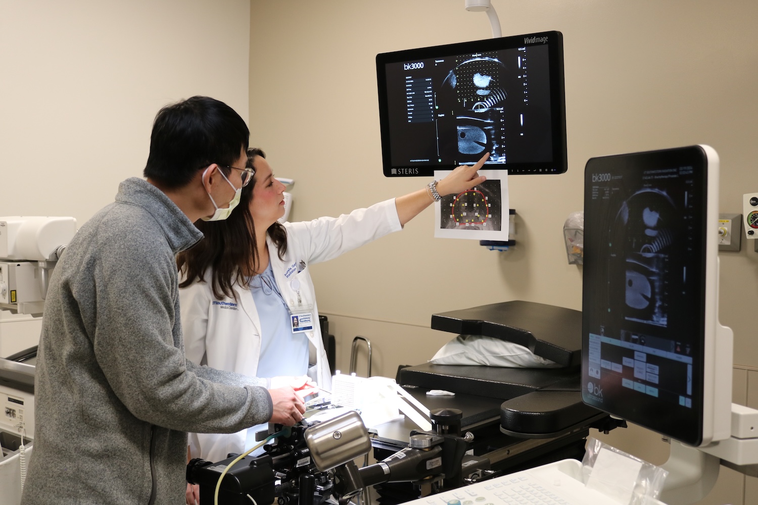 Two individuals in a clinical setting review medical imaging scans on dual monitors; one, wearing a lab coat and ID badge, points at the screen while the other observes.
