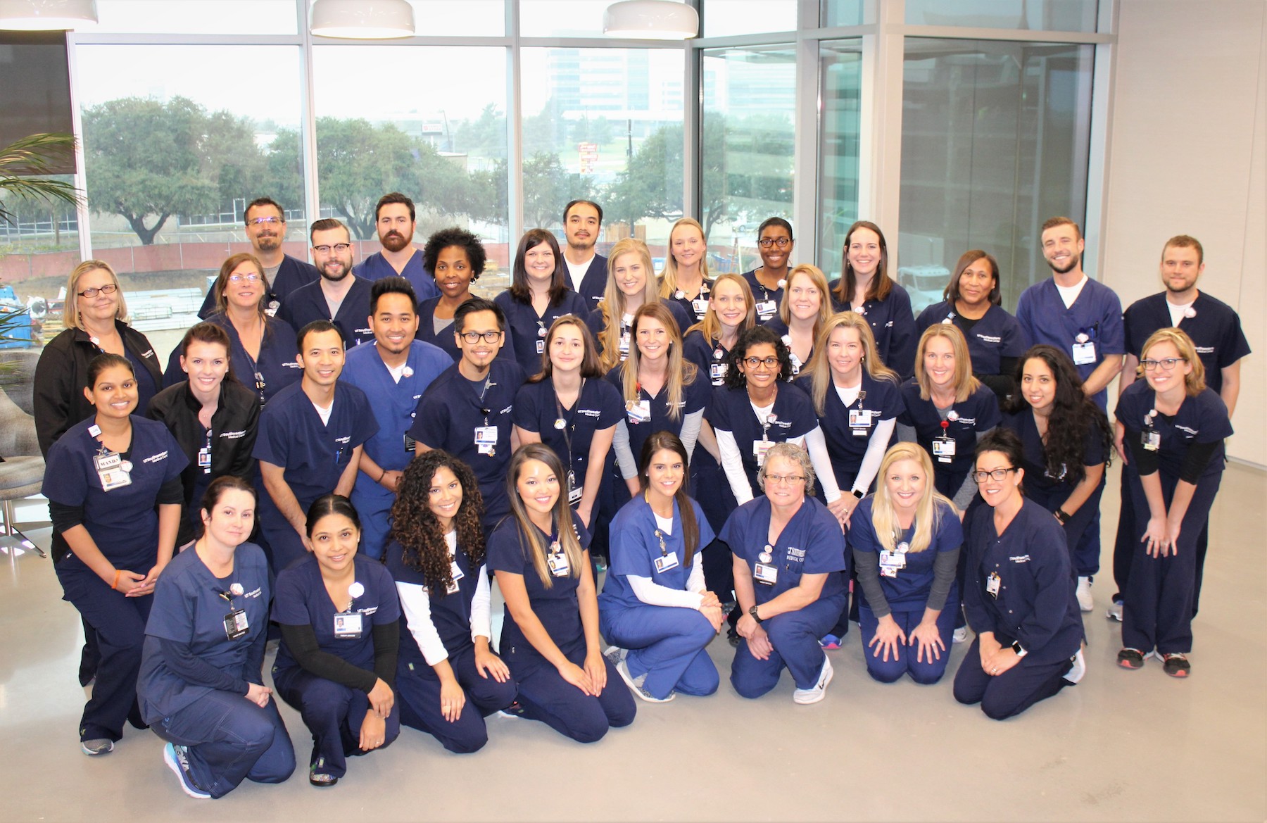 Group photo of healthcare professionals in navy blue scrubs with ID badges, taken indoors in a building with large windows in the background.