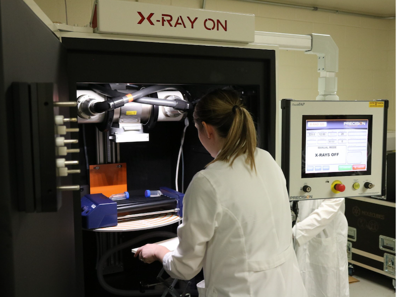 An individual in a white lab coat operates an X-RAD 320 machine. The individual is adjusting equipment inside the machine, while a nearby monitor displays various settings and shows 'X-RAYS OFF.