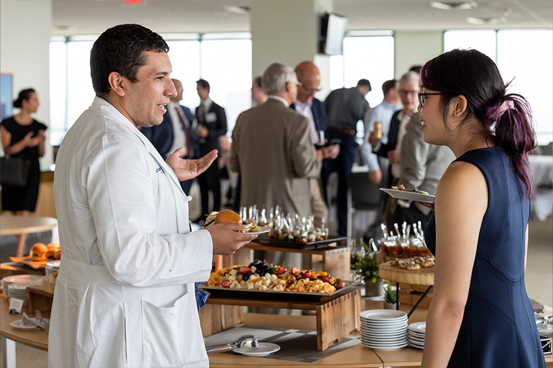 Man in white coat talking to woman in navy dress while holding plates of food