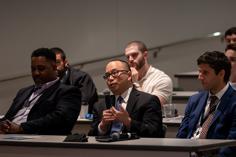 Man with glasses and black suit holding microphone in audience and speaking