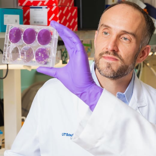 male student in white lab coat and purple gloves looks at petri dish