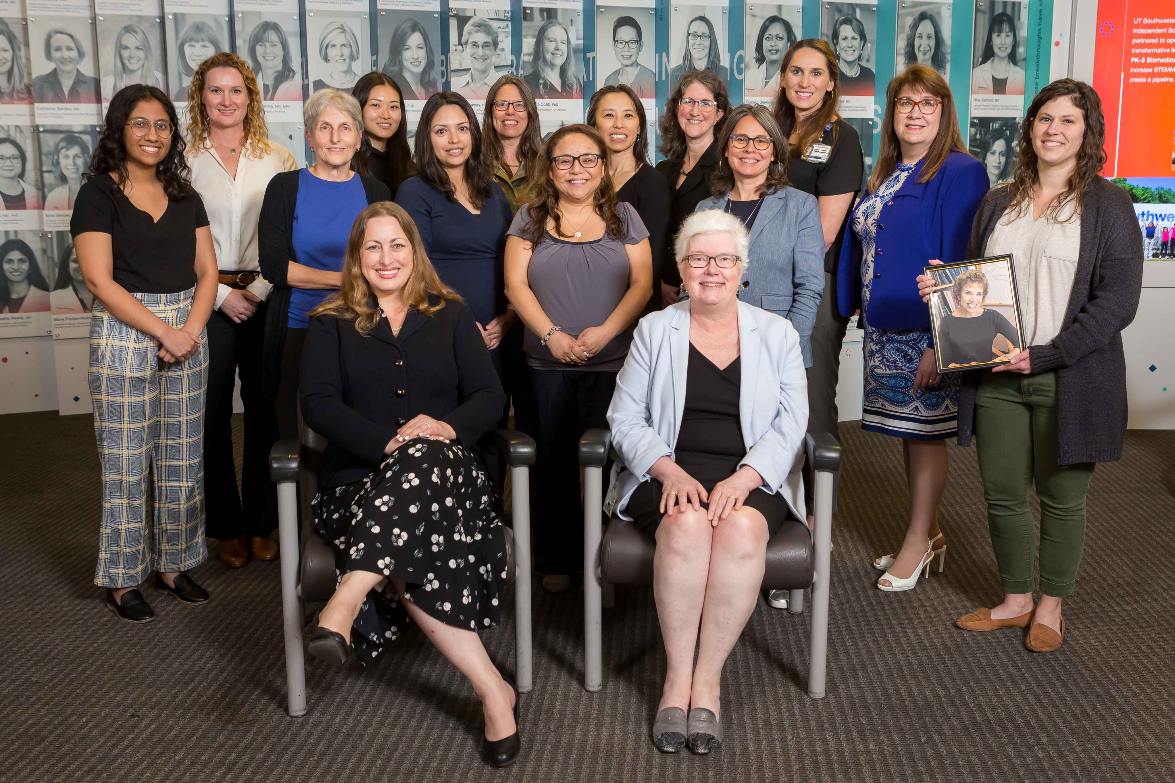 A group photo. A woman standing at the right of the photo is holding a portrait of Dr. Carole Mendelson