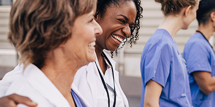 2 smiling women in white coats and blue scrubs in foreground, 2 people in blue scrubs in background