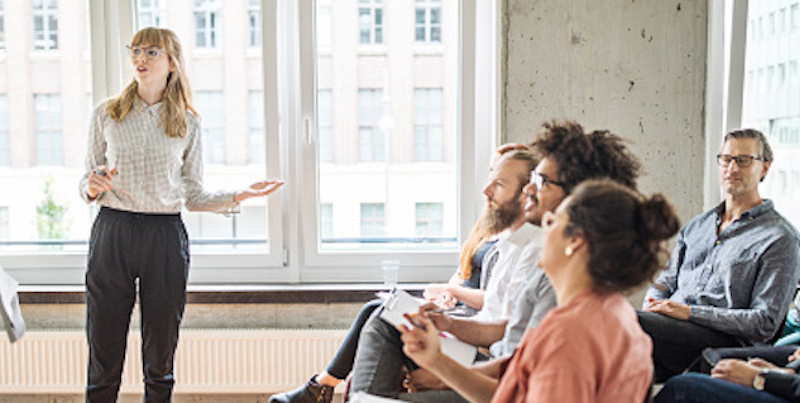 woman pointing at white board notes while standing in front of class of seated adult students