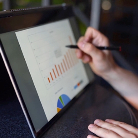 laptop screen displays colorful bar chart with person's hand and pen pointing at screen
