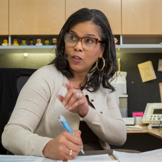 Close up of female sitting at desk with a pen in her float-right hand. She is looking at and speaking with someone outside of the camera frame.