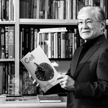 Dr. Takahashi holding a brochure, standing in front of a library full of books