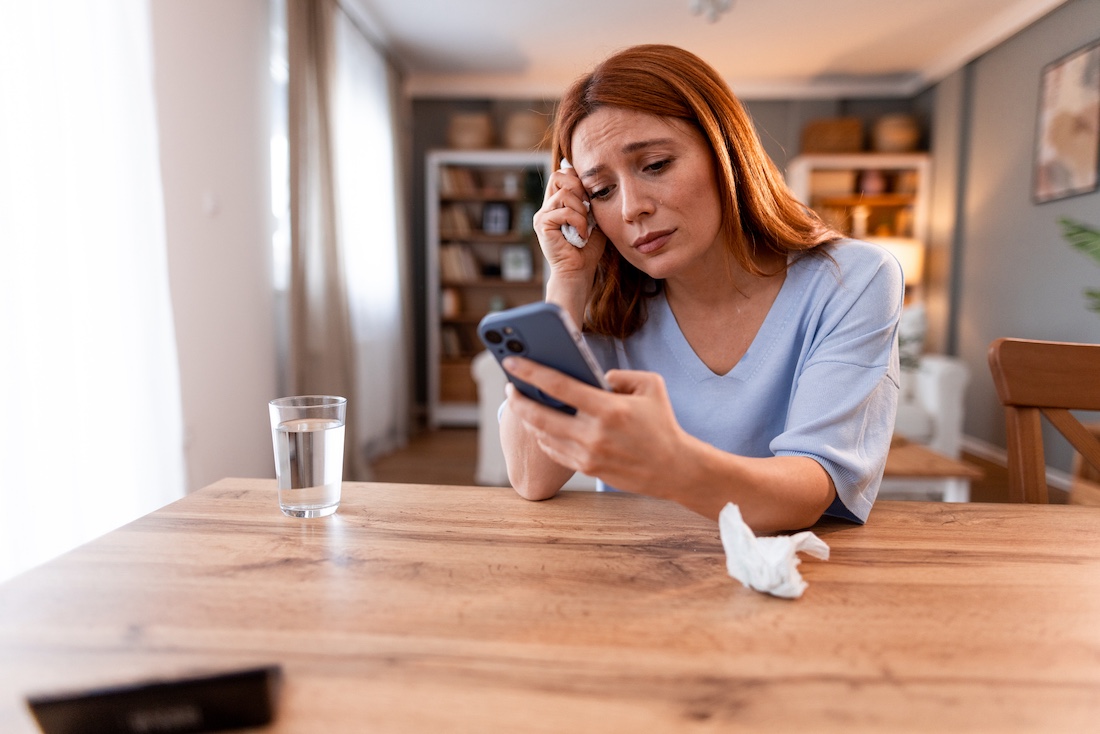 A worried woman sitting at a table looking at her phone with a thoughtful expression. 