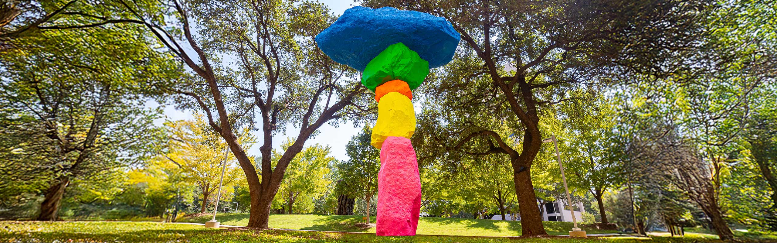 A photo of Ugo Rondinone&rsquo;s Dallas Mountain. The sculpture is made up of colorfully painted boulders, with the largest one on top. It is placed in a green part of campus, surrounded by trees.