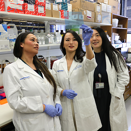 Three young women in white lab coats looking at a computer screen as the woman in the middle points at data.