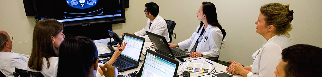 Doctors sitting at a meeting room table with laptops in front of them and a flatscreen on the wall.